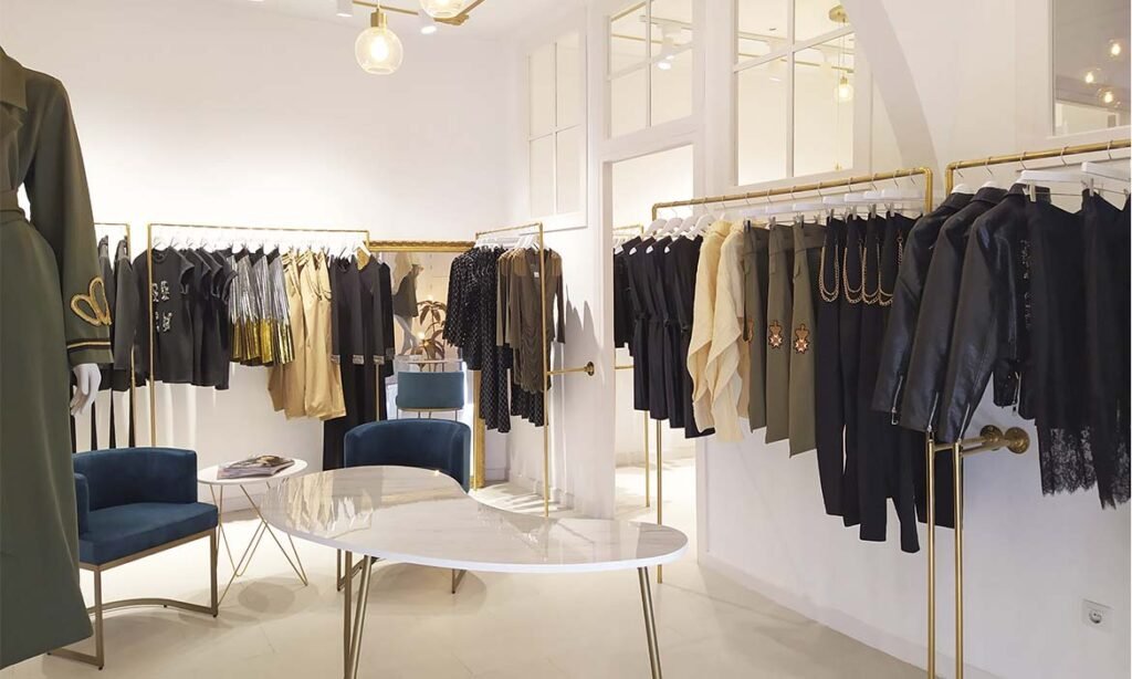 Tendances Boutique de mode haut de gamme avec vêtements de luxe exposés sur des portants dorés. Collection exclusive de tenues élégantes en noir, doré, beige et vert, ornées de broderies et de détails métalliques. Décoration raffinée avec une table en marbre et des fauteuils en velours bleu. Espace lumineux et design moderne, idéal pour une expérience shopping premium Tendance
